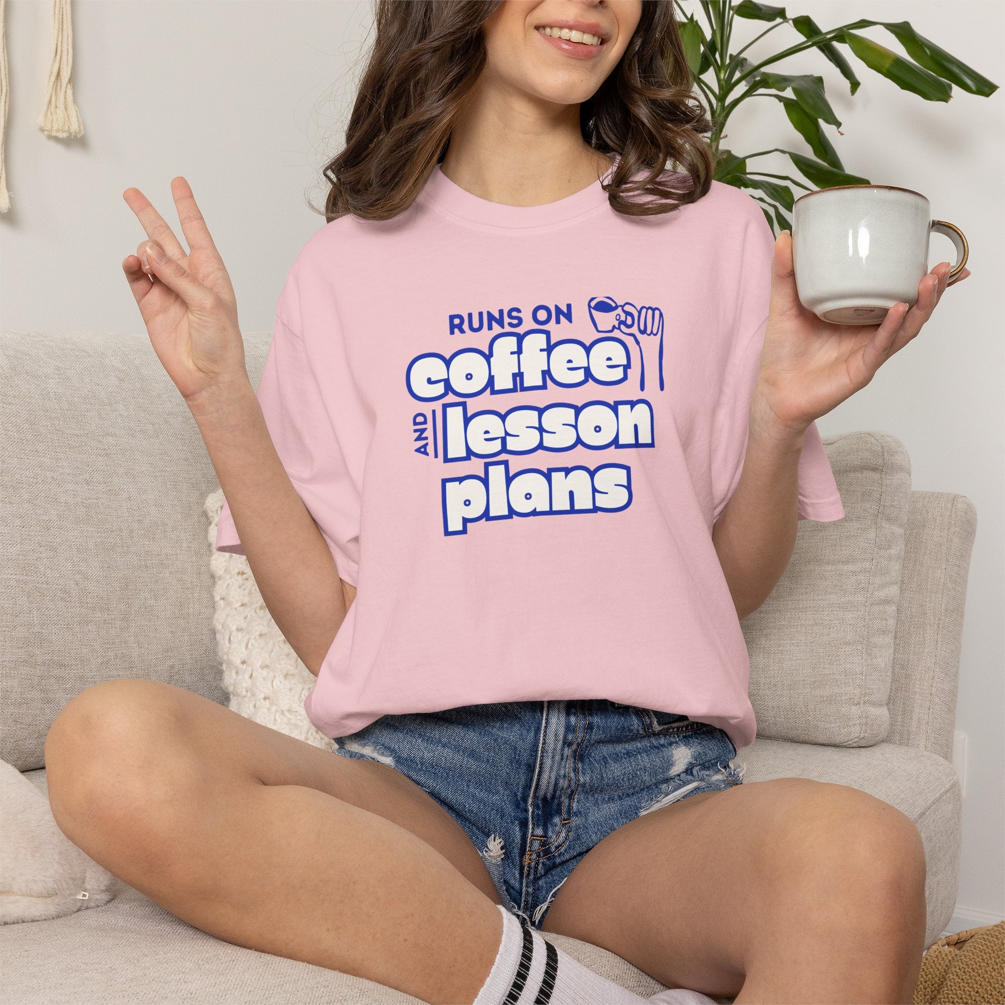 A woman sitting on a light beige couch wearing a lavender crewneck t-shirt with a colorful graphic that reads 'OCCUPATIONAL THERAPY' in rainbow letters around a smiley face. She is holding a white ceramic mug.