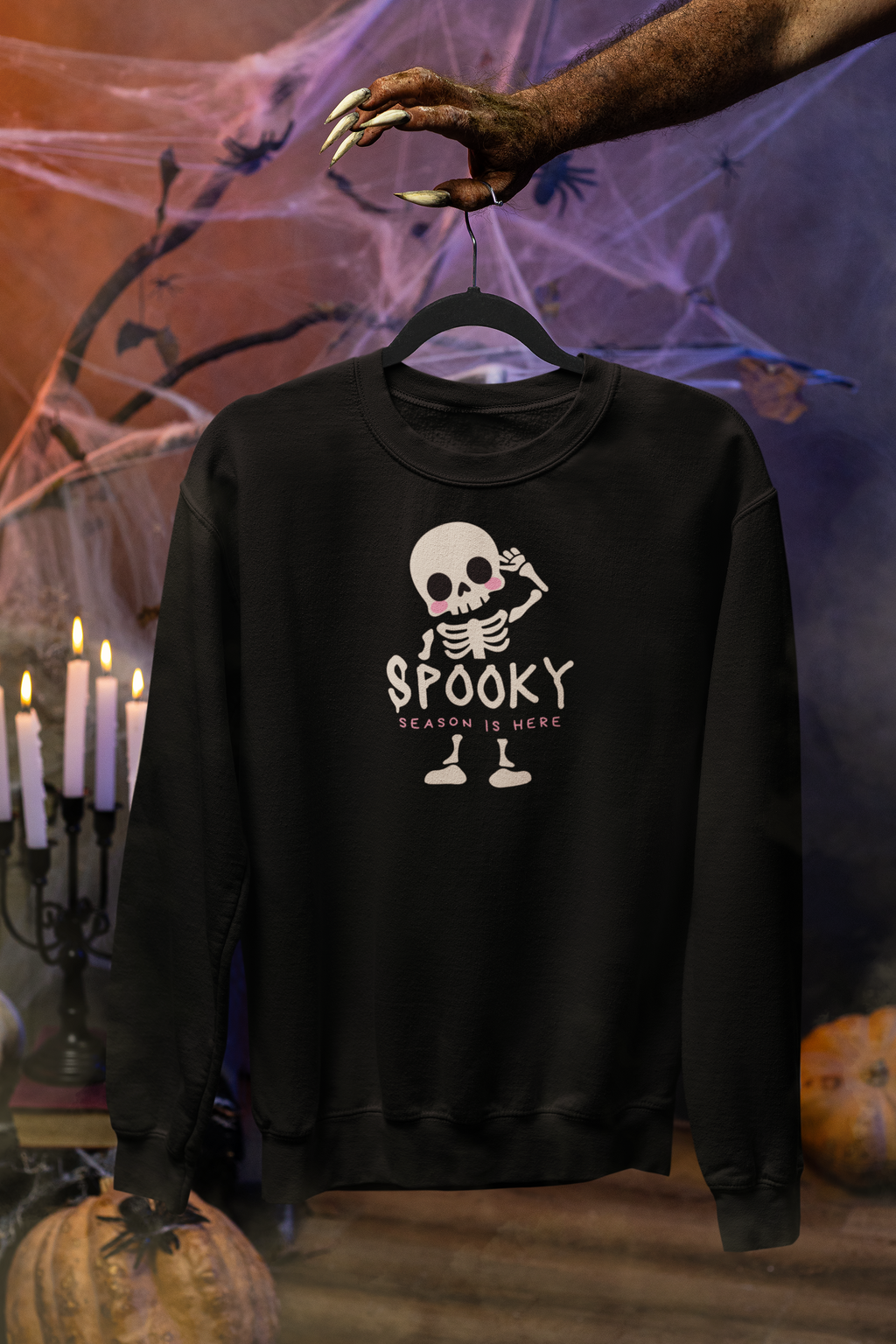 A black crewneck sweatshirt hanging on a hanger, featuring a cartoon skeleton graphic with the text 'SPOOKY SEASON IS HERE'. The background is a Halloween-themed set with a hand holding the hanger, spiderwebs, candles, and pumpkins.