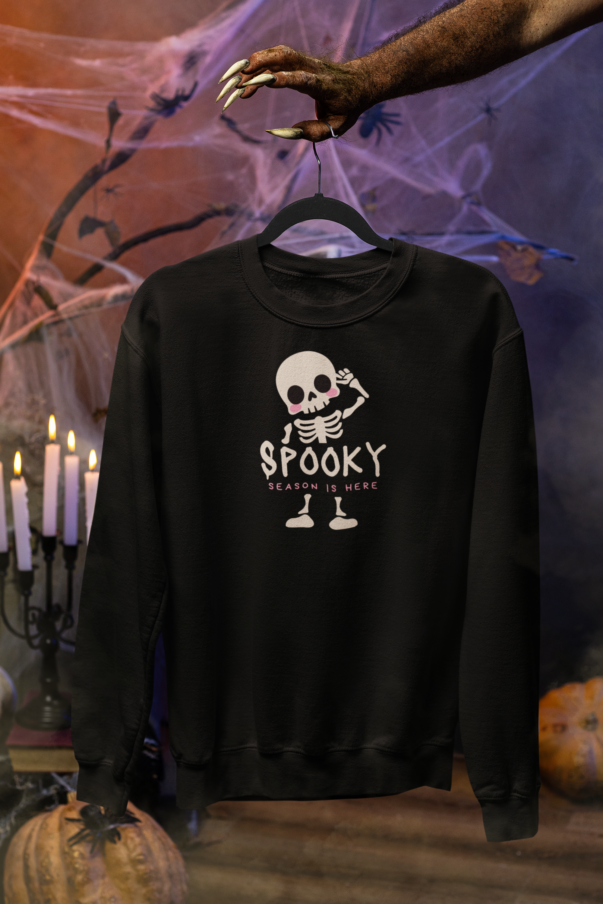 A black crewneck sweatshirt hanging on a hanger, featuring a cartoon skeleton graphic with the text 'SPOOKY SEASON IS HERE'. The background is a Halloween-themed set with a hand holding the hanger, spiderwebs, candles, and pumpkins.