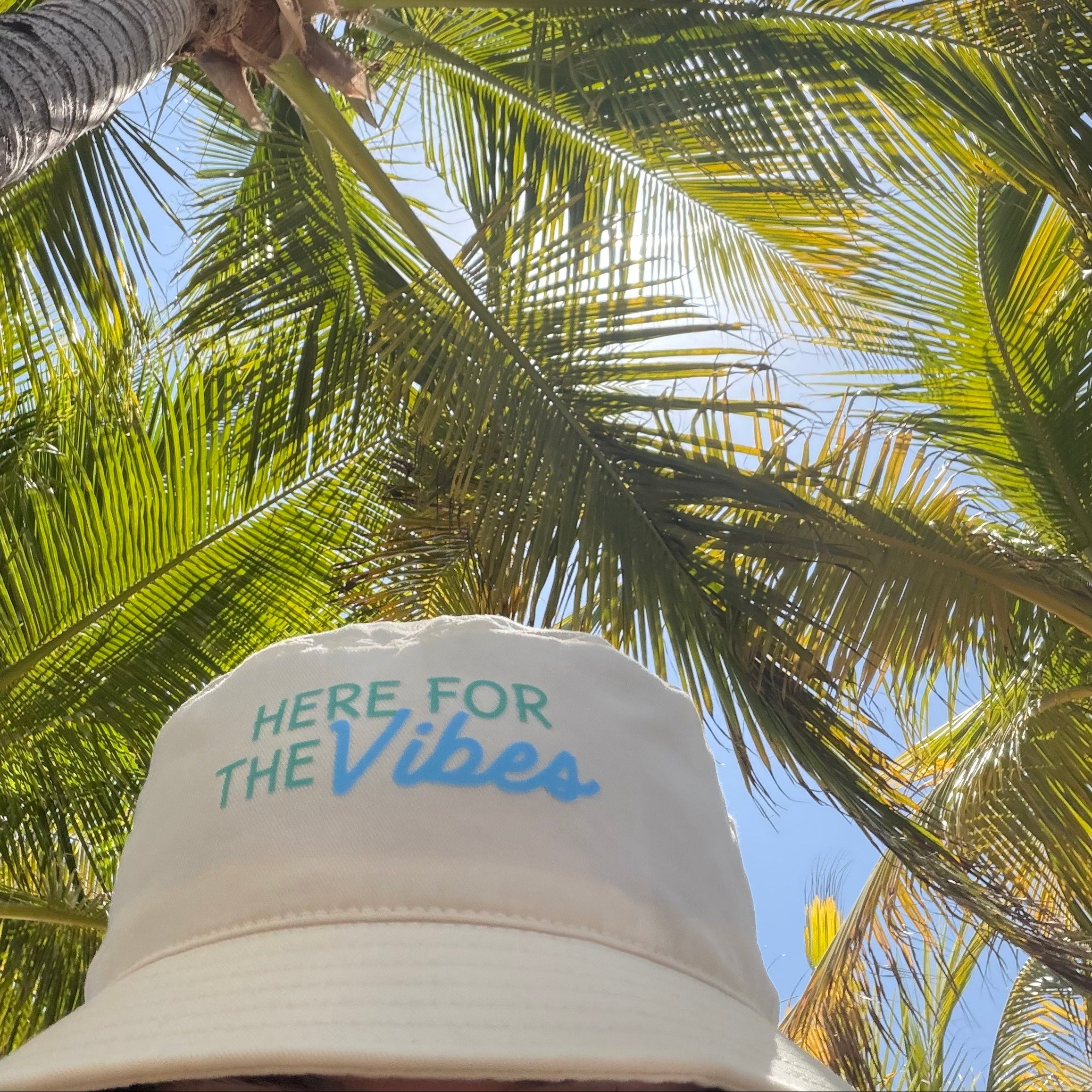 A person wearing a beige bucket hat with 'HERE FOR THE Vibes' embroidered on the front, photographed from below among green palm leaves and blue sky.