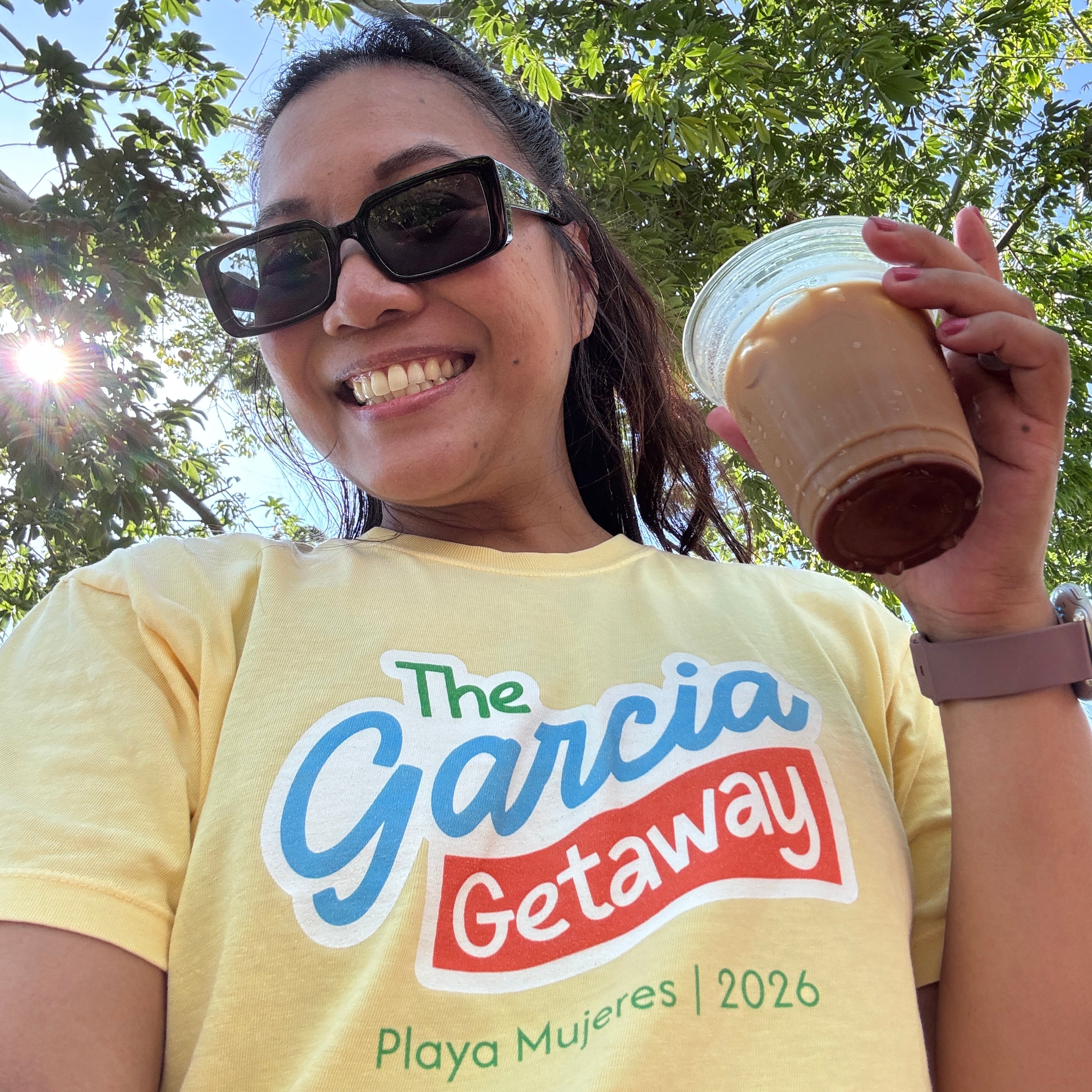 A smiling woman outdoors under leafy trees wears a yellow graphic T-shirt that reads 'The Garcia Getaway Playa Mujeres | 2026' and holds a clear plastic cup of iced coffee.